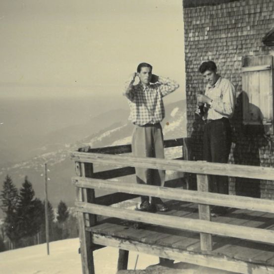 Ein Schwarz-Weiß-Foto von zwei Männern auf einem Balkon in einem verschneiten Berggebiet. Ein Mann berührt seine Haare, der andere hält eine Flasche. Im Hintergrund sind Bäume und ein Pfahl zu sehen.
