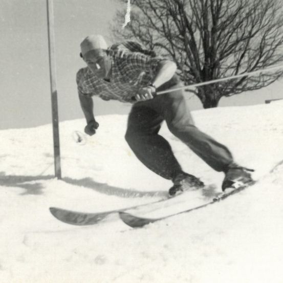 Ein Schwarz-Weiß-Foto eines Mannes, der mit einer Zigarette im Mund und einem Baum im Hintergrund bergab Ski fährt.