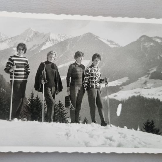Ein Schwarz-Weiß-Foto zeigt vier Frauen im Schnee, die Skistöcke halten. Dahinter sind Berge und Bäume zu sehen.