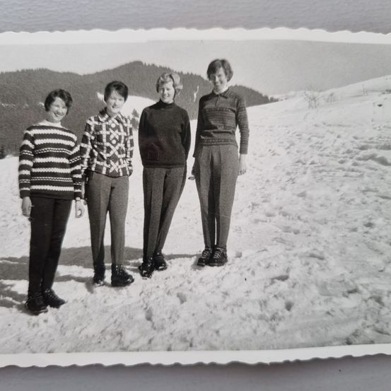 Vier Frauen posieren für ein Foto auf einem verschneiten Berg. Sie alle lächeln und tragen warme Kleidung. Im Hintergrund befindet sich eine bergige Landschaft.