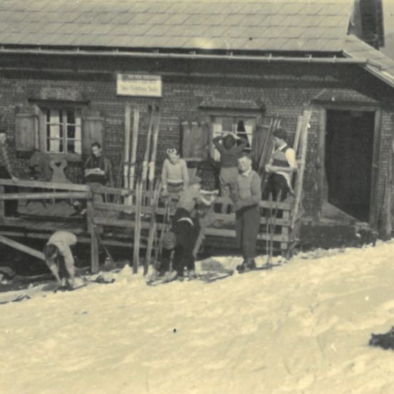 Ein Schwarz-Weiß-Foto zeigt eine Gruppe von Menschen vor einem Backsteingebäude, einige stehen auf Skiern und andere auf einem Holzzaun. Skistöcke sind gegen die Wand gelehnt.