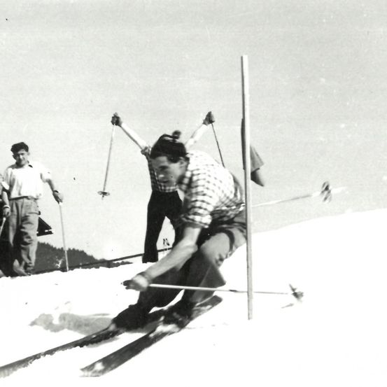 Ein Skifahrer fährt bergab und umgeht Stangen, während zwei Männer hinter ihm auf einem verschneiten Hang stehen.