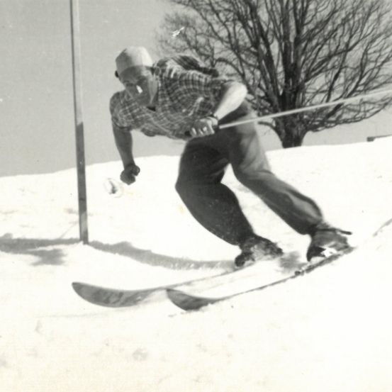 Ein Schwarz-Weiß-Foto eines Mannes, der eine verschneite Hügel hinunterskit. Er trägt eine Mütze und ein kariertes Hemd. Ein Baum ist im Hintergrund sichtbar.