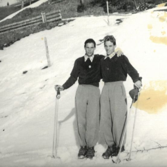 Zwei Männer stehen im Schnee mit Skistöcken, einer hat seinen Arm um die Schulter des anderen gelegt.