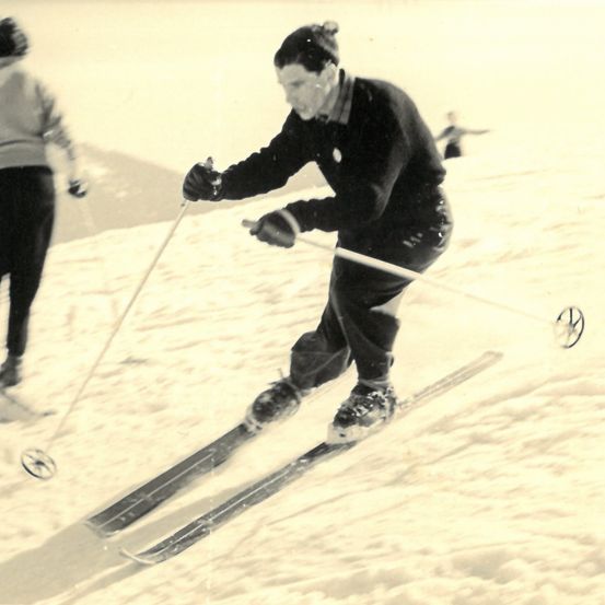 Ein Schwarz-Weiß-Foto zeigt einen Mann, der mit Skistöcken den Schneehang hinunterskit, einen Pullover und Handschuhe trägt. Ein weiterer Skifahrer ist im Hintergrund zu sehen, der ebenfalls Ski fährt.