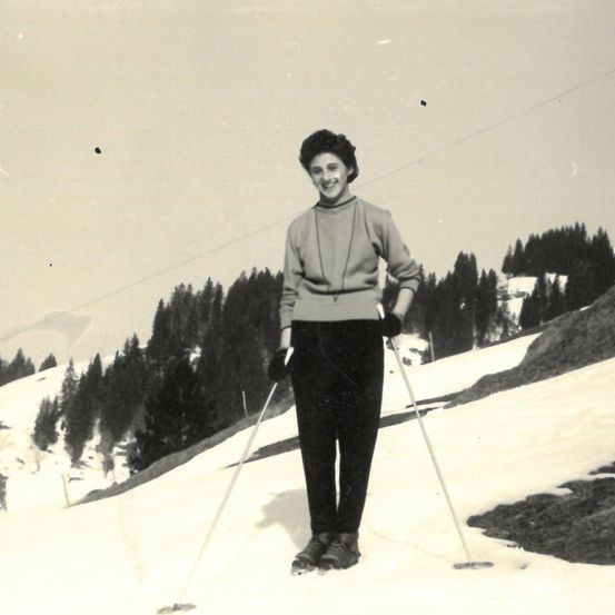 Ein altes Schwarz-Weiß-Foto zeigt eine Frau auf einem verschneiten Hügel mit Skiern. Sie lächelt und ist in warme Winterkleidung gekleidet. Bäume und Berge sind im Hintergrund zu sehen.