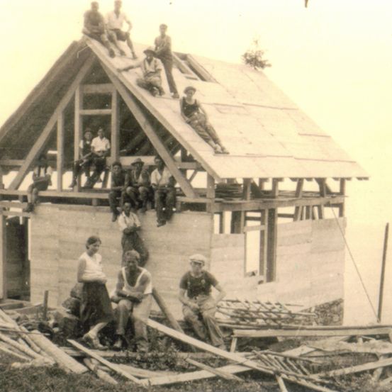 Eine Gruppe von Menschen arbeitet an einer Holzkonstruktion. Einige sind auf dem Dach, während andere auf dem Boden sitzen. Einige halten Werkzeuge.