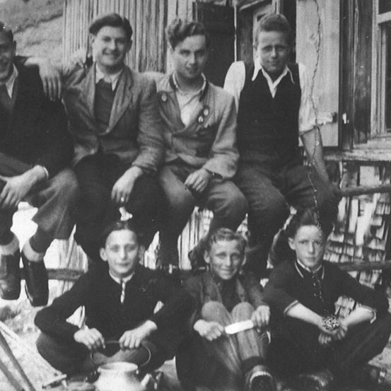 Eine Gruppe junger Männer in Anzügen und Krawatten posiert vor einem Holzgebäude. Einige sitzen, andere stehen. Sie scheinen für ein Foto zu posieren.