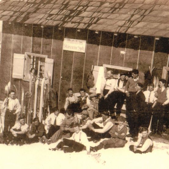 Ein altes Foto zeigt eine Gruppe von Männern in Winterkleidung, die vor einem Gebäude mit einem Schild 'Starvin's' sitzen und stehen. Einige halten Skier und andere stehen an der Wand.