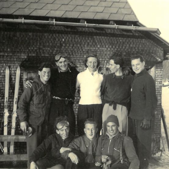 Ein Schwarz-Weiß-Foto von sieben Personen vor einem Haus mit Ziegelwänden und einem Dach. Zwei Personen sitzen in der vorderen Reihe. Sie sind alle für kaltes Wetter gekleidet.