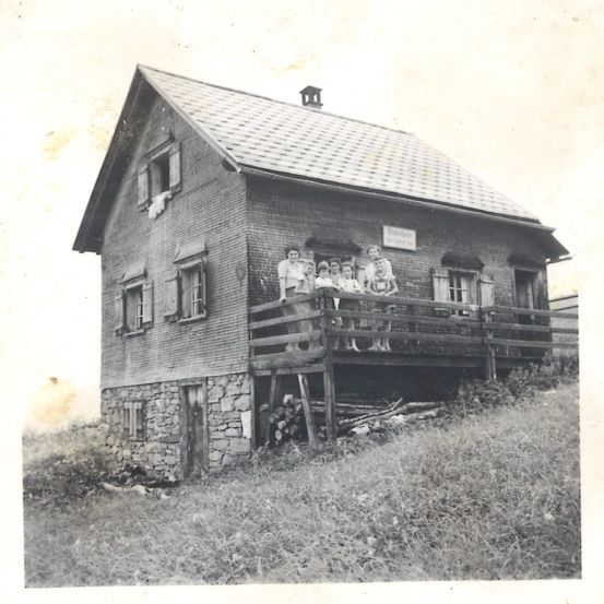 Ein altes Schwarz-Weiß-Foto zeigt eine Gruppe von Menschen auf der Veranda eines Backsteinhauses mit Schornstein.