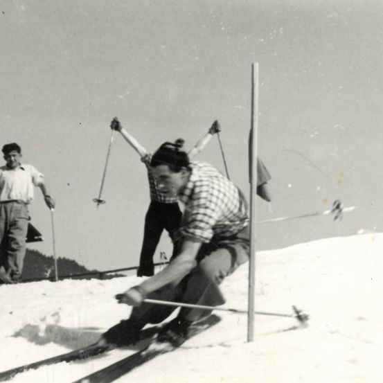 Ein Schwarz-Weiß-Foto von drei Menschen, die auf einem verschneiten Hang Ski fahren. Ein Skifahrer ist im Vordergrund und fährt bergab. Dahinter halten zwei andere Stangen hoch.