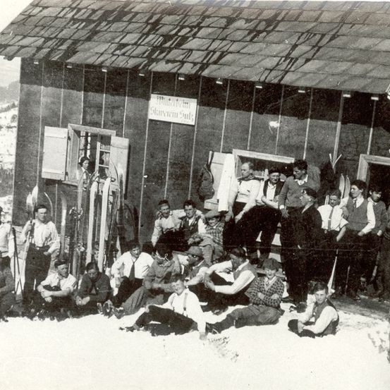 Eine Gruppe von Menschen, gekleidet in Vintage-Winterkleidung, posiert vor einem Gebäude mit dem Schild 'Schanzerhaus'. Die Umgebung ist verschneit, und die Personen sitzen und stehen in verschiedenen Posen.