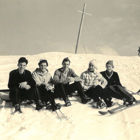 Fünf Personen sitzen im Schnee, lächeln und posieren für ein Foto. Sie tragen Winterkleidung, Mützen und halten Skistöcke. Der Schnee ist mit einer dicken Schneeschicht bedeckt. Ein Holzpfahl steht hinter ihnen.