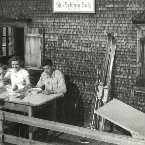 Ein Schwarz-Weiß-Foto zeigt zwei Personen, die an einem Tisch sitzen, möglicherweise in einem Chalet. Ein Schild darüber lautet 'Shi-Schnion Sub'. Skier sind an der Wand gelehnt.