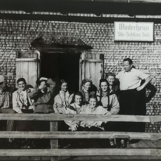 Ein Schwarz-Weiß-Foto einer Gruppe von Menschen, die vor einem hölzernen Gebäude stehen und sitzen. Sie sind in altmodischer Kleidung gekleidet. Das Gebäude hat ein Schild, das Winterheim Ski-Sektion Sulz besagt.