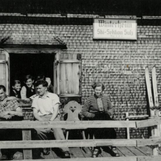 Ein Schwarz-Weiß-Bild einer Gruppe von Menschen, die auf einer Holzbank vor einem Gebäude namens Winterheim sitzen.