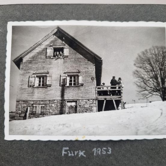 Schwarz-Weiß-Foto eines Hauses mit Menschen auf einem Balkon und anderen, die auf einer Veranda davor stehen, datiert 1953, umgeben von Schnee.