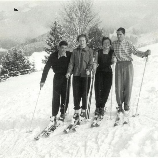 Vier Personen, in Winterkleidung gekleidet, stehen auf einem verschneiten Hügel und lächeln für die Kamera. Dahinter ragen immergrüne Bäume in die Landschaft.