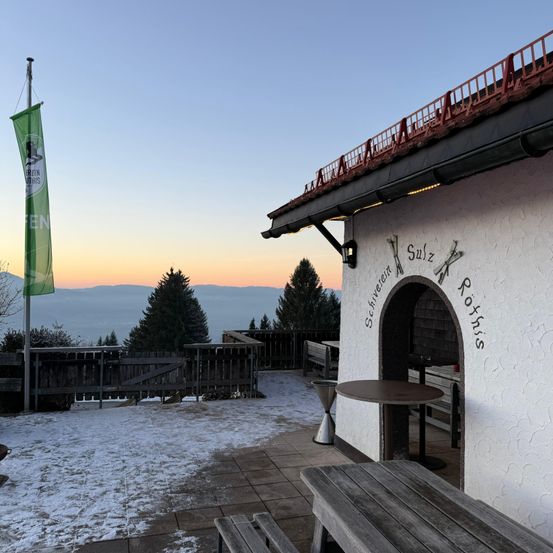 Ein Chalet mit roten Geländern und einer Fahne steht in einer verschneiten Landschaft. Ein Eingangsbereich trägt Sulz Rottis. Bänke und ein Tisch stehen draußen.