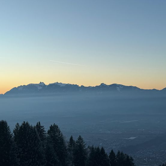Sonnenuntergang über einer Bergkette mit einem Wald im Vordergrund und einer Stadt im Hintergrund. Der Himmel ist orange und blau.