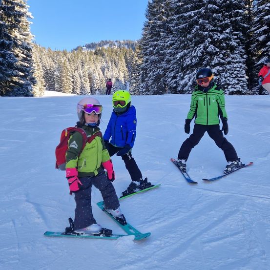 Drei Kinder fahren auf einem verschneiten Hang mit Tannen im Hintergrund. Jedes Kind trägt farbenfrohe Skiausrüstung. Das Mädchen links trägt einen Rucksack.