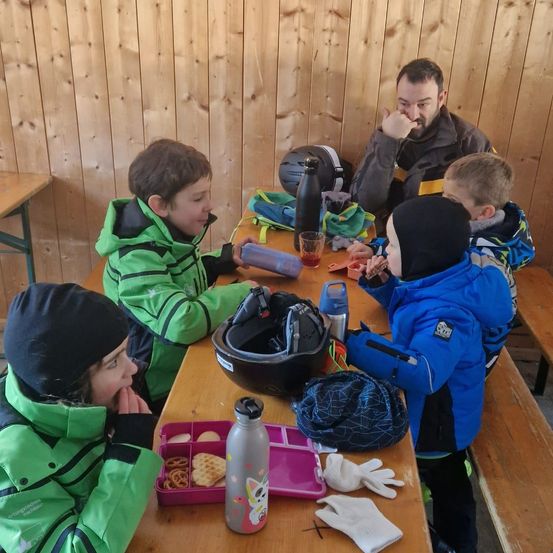 Eine Familie sitzt an einem Tisch in einem Holzraum. Ein Mann und vier Kinder essen Snacks. Auf dem Tisch sind ein Helm, Becher, eine Flasche und Handschuhe. Die Wand ist aus Holz.