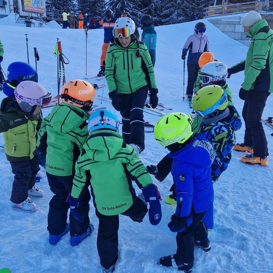 Eine Gruppe von Kindern in grünen und blauen Winterjacken steht auf einem verschneiten Hang. Sie sind von Erwachsenen in Skiausrüstung umgeben. Die Kinder tragen Helme, Handschuhe und Skischuhe. Ein Erwachsener hält einen Skistock.