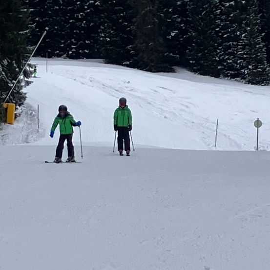 Zwei Personen sind auf einem verschneiten Hang beim Skifahren, tragen grüne Jacken, Handschuhe und Helme. Eine ist etwas voraus. Im Hintergrund sind Kiefernbäume zu sehen.