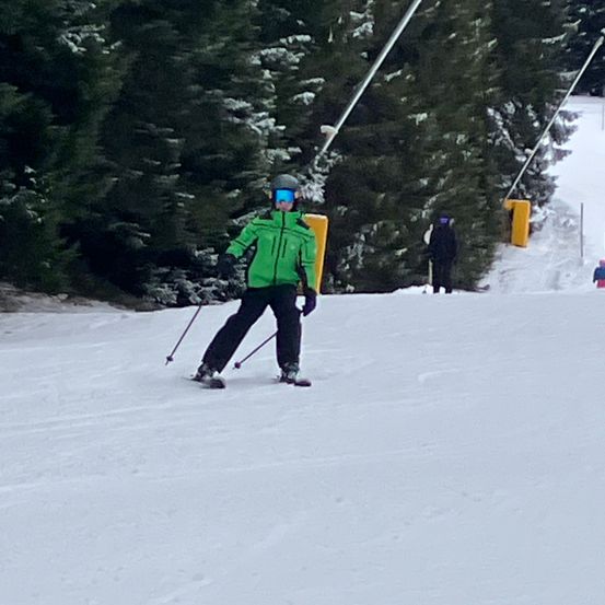Eine Person, die einen verschneiten Hang hinunterskit, mit Tannenbäumen und Skiliftstangen im Hintergrund.