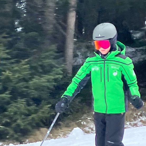 Ein Skifahrer in einer grünen Jacke, schwarzen Hosen und roten Brille steht auf einem verschneiten Hang mit Tannen im Hintergrund.
