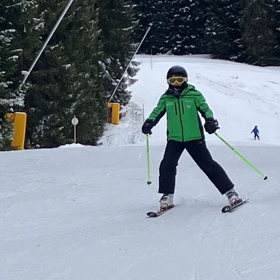Ein Skifahrer in einer grünen Jacke und einer Schutzbrille fährt einen verschneiten Hang hinunter, mit Tannen und Stangen im Hintergrund. Ein weiterer Skifahrer ist in der Ferne sichtbar.