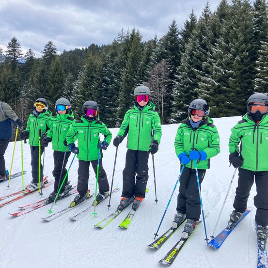 Eine Gruppe von Skifahrern in grünen Jacken und Helmen posiert für ein Foto auf einer verschneiten Piste. Sie sind von schneebedeckten Bäumen umgeben.