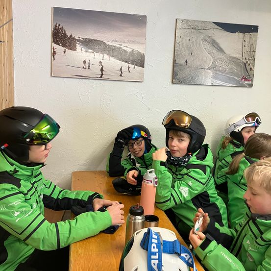 Eine Gruppe von Kindern in grünen Skijacken sitzt an einem Tisch, einige tragen Helme. Sie haben Skiausrüstung um sich herum, einschließlich Helme und eine Wasserflasche. Die Wand hinter ihnen hat zwei Poster mit verschneiten Landschaften.