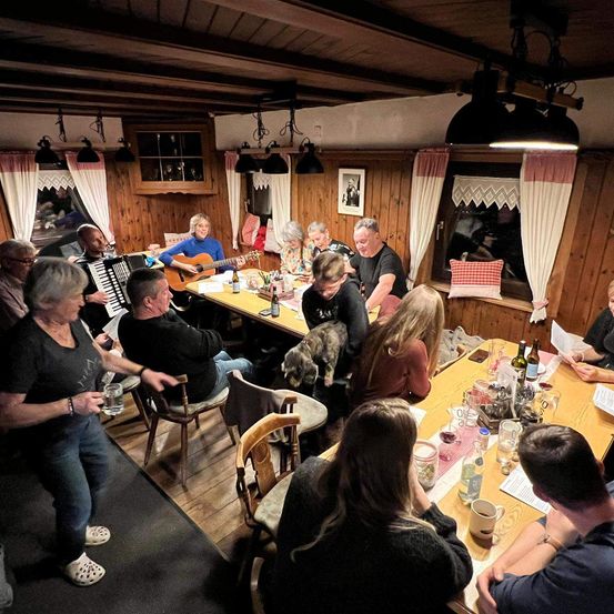 Eine Gruppe von Menschen versammelt sich in einem Raum mit Holzwänden, Tischen und Stühlen. Einige sitzen, andere stehen, und eine Frau spielt Akkordeon. Flaschen, Gläser und eine Katze sind auf dem Tisch. Es gibt Lampen und Bilderrahmen an der Wand.