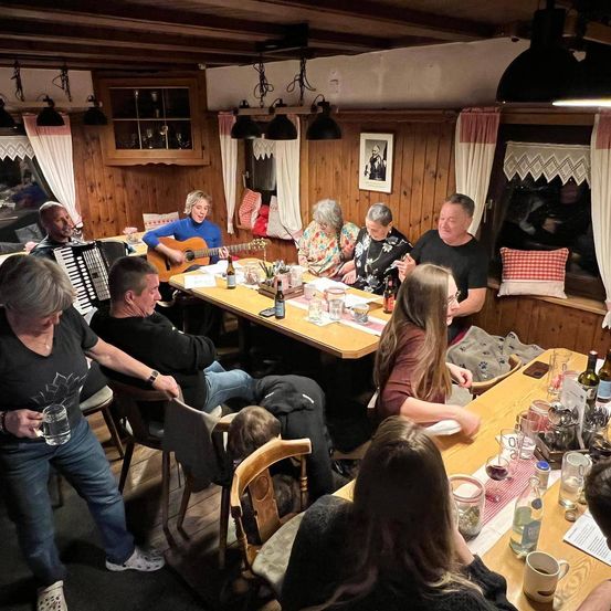 Eine Gruppe Erwachsener versammelt sich um einen Tisch in einem hölzernen Raum. Einige spielen Musikinstrumente, während andere zusehen. Flaschen, Gläser und ein Baby sind auf dem Tisch.