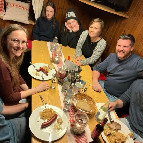 Sechs Personen sitzen um einen Holztisch in einem rustikalen Ambiente und genießen ihre Mahlzeit. Zwei Frauen tragen Brillen, und eine von ihnen lächelt fröhlich. Der Tisch ist mit Tellern, Gabeln, Messern und verschiedenen Getränken besetzt. Dahinter befindet sich ein Regal mit Fernseher und dekorativen Kissen.