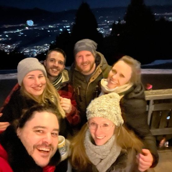 Fünf Personen lächeln und posieren für ein Foto mit einer nächtlichen Stadtsilhouette im Hintergrund. Sie tragen alle Winterkleidung.