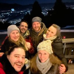 Fünf Personen lächeln und posieren für ein Foto mit einer nächtlichen Stadtsilhouette im Hintergrund. Sie tragen alle Winterkleidung.