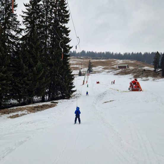 Ein Kind in einer blauen Jacke fährt einen schneebedeckten Hang hinunter, mit einer Skiliftstation im Hintergrund.