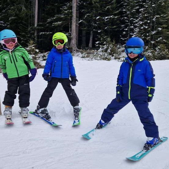 Drei Kinder stehen Seite an Seite im Schnee und machen Ski. Sie tragen Winterkleidung und Skiausrüstung. Bäume umgeben sie, und der Schnee ist dick.