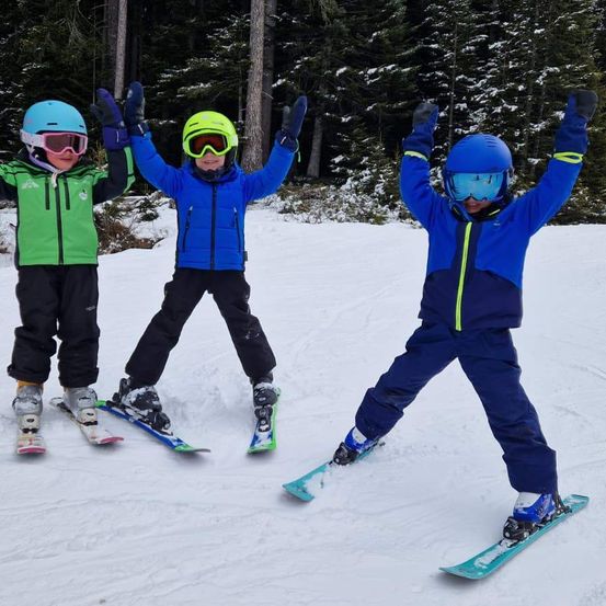 Drei Kinder, alle mit Skiausrüstung, stehen auf einer verschneiten Piste. Sie haben ihre Arme zur Feier erhoben. Im Hintergrund befinden sich Bäume mit Schnee auf ihren Ästen.
