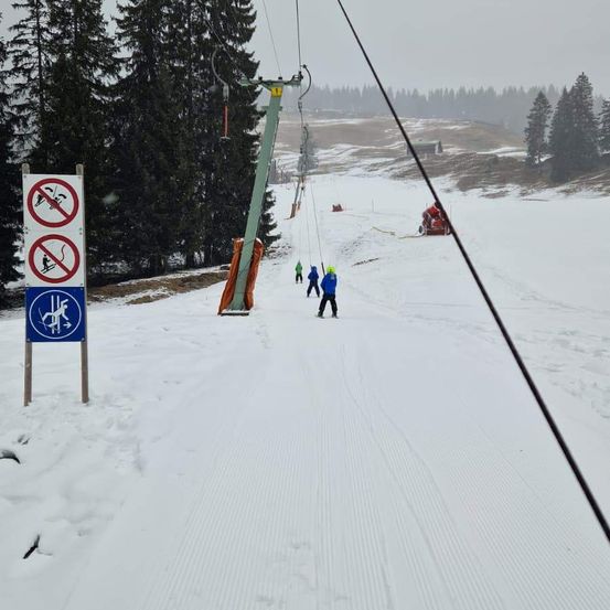 Eine verschneite Skipiste mit einem umgestürzten Skilift und Skifahrern. Bäume und ein Gebäude sind im Hintergrund.