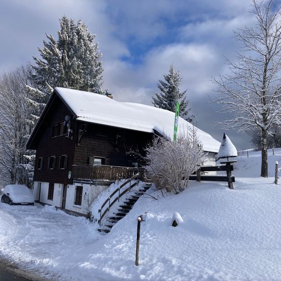 Ein von Schnee bedecktes Chalet mit einem davor geparkten Auto. Bäume und ein Zaun umgeben das Haus, wobei eine Fahne in der verschneiten Landschaft sichtbar ist.