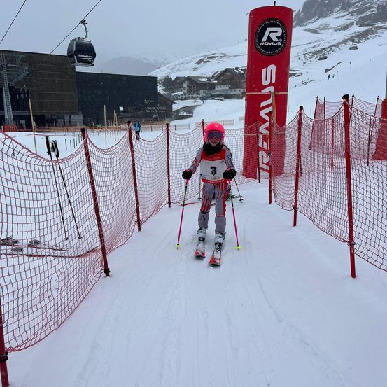Ein Skifahrer in einem rosa Helm und einer Schutzbrille navigiert einen rot eingenetzten Kurs auf einem verschneiten Hang, mit einem großen roten Banner, das 'Remus' im Hintergrund liest.