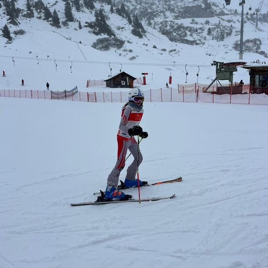 Ein Skifahrer mit Helm und Brille fährt einen verschneiten Hang hinunter. Der Skifahrer hält Skistöcke und trägt Skischuhe. Im Hintergrund befinden sich Skilifte und ein kleines Gebäude.