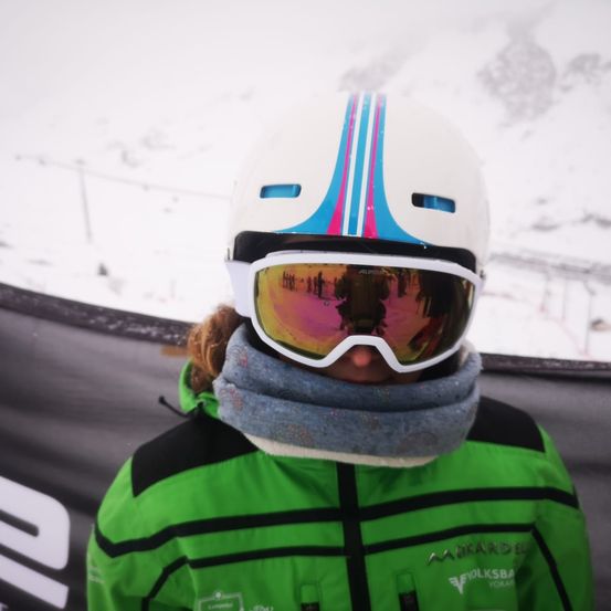 Ein Skifahrer mit einem weißen Helm mit blauen und rosa Streifen, reflektierenden Brillengläsern und einem grauen Schal steht vor einem verschneiten Berg.