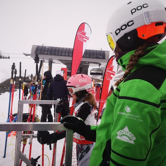 Ein Skifahrer mit einem rosa Helm und Schutzbrille steht neben einer Person in einer grünen Jacke an einem Skilift. Viele Skistöcke sind sichtbar, und es gibt rote Banner mit weißem Text im Hintergrund.