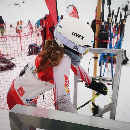 Ein Skifahrer bereitet sich darauf vor, einen verschneiten Hang hinabzufahren, ausgestattet mit einem weißen Helm, einer Brille und einem roten und weißen Skianzug. Hinter ihr sind mehrere Personen sichtbar, einige tragen Skiausrüstung und andere stehen in der Nähe von Skistöcken.