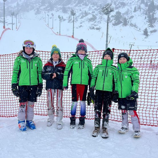 Fünf junge Personen in grüner und weißer Skiausrüstung posieren für ein Foto im Schnee, vor einem roten Netz stehend.